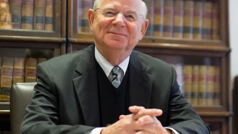 Elderly man in a suit sits at a desk with law books behind him, smiling slightly.