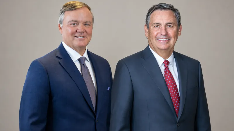 Two men in suits smiling, one in blue and one in gray with a red tie.