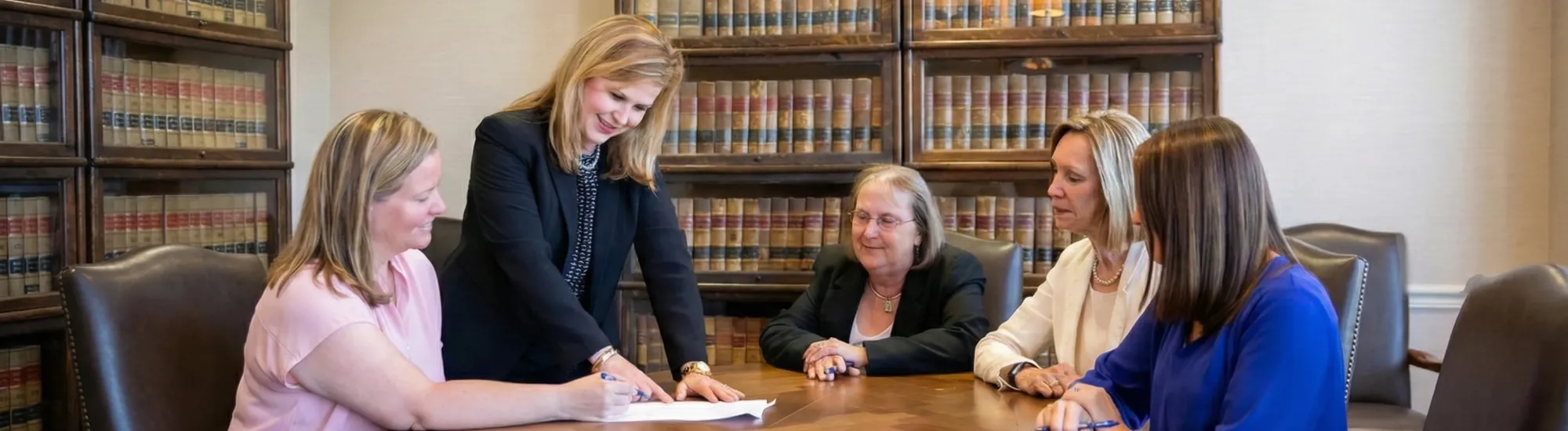 Five women in a library meeting room discuss documents at a large wooden table.